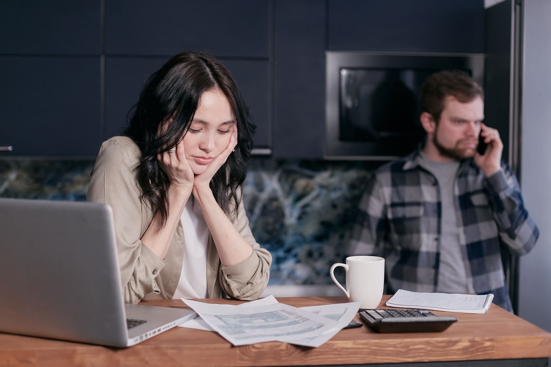 person ved køkkenbord med bærbar computer og elregning, kuvert og kaffekop, dagslys fra vindue, nordisk hjem